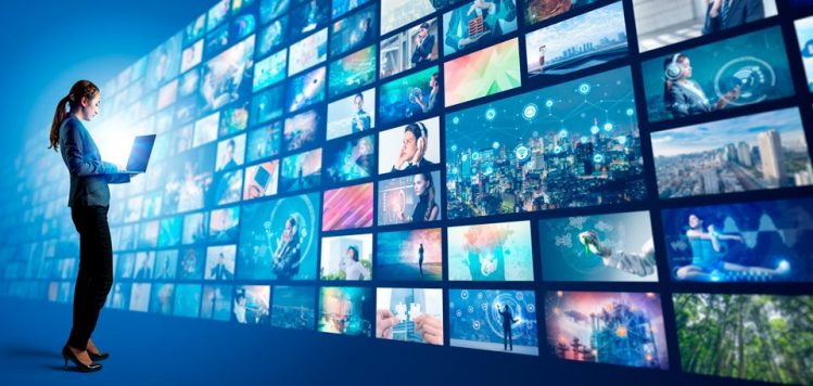 Woman with a laptop in front of a large wall of digital video screens